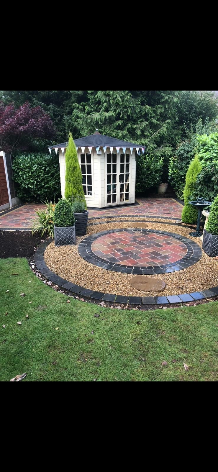 Garden with round brick patio and small white gazebo surrounded by plants and trees.