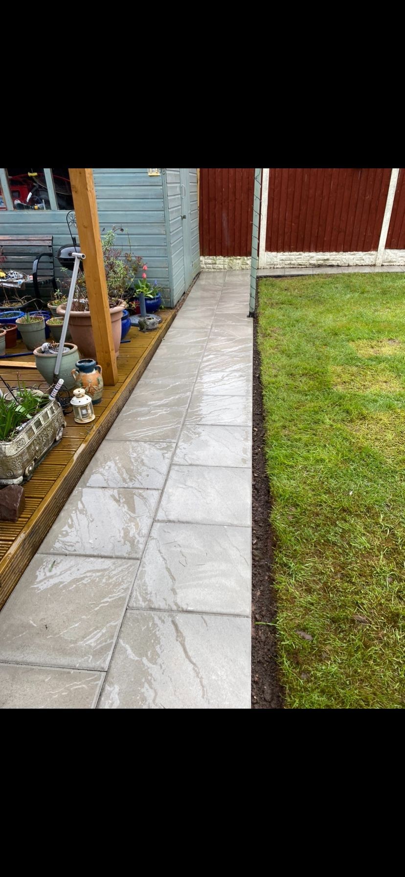 Garden path with gray tiles alongside a wooden deck with potted plants and lawn area.