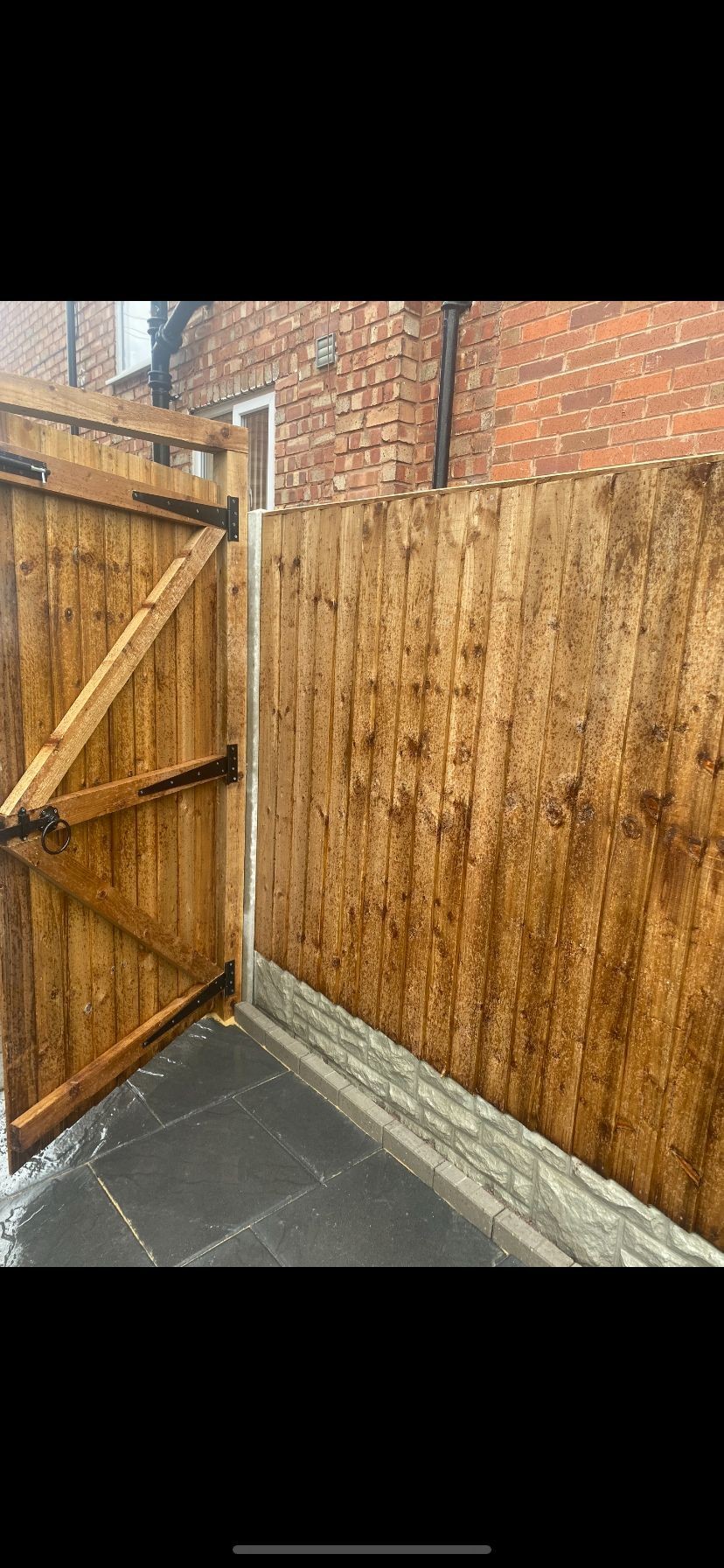 Wooden gate and fence along a brick house with a stone base on a dark tiled patio.