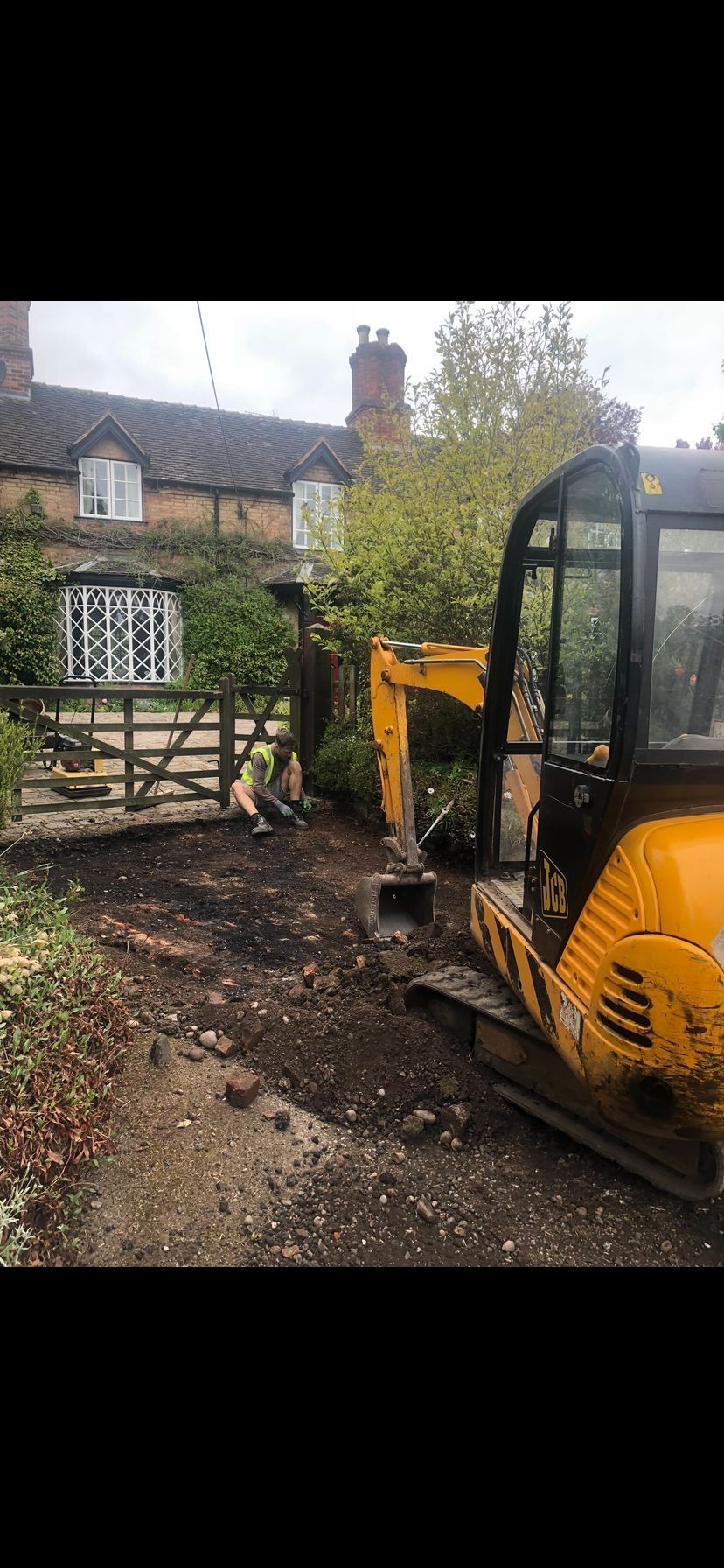 Construction site with a yellow excavator and a worker in a safety vest near a cottage.