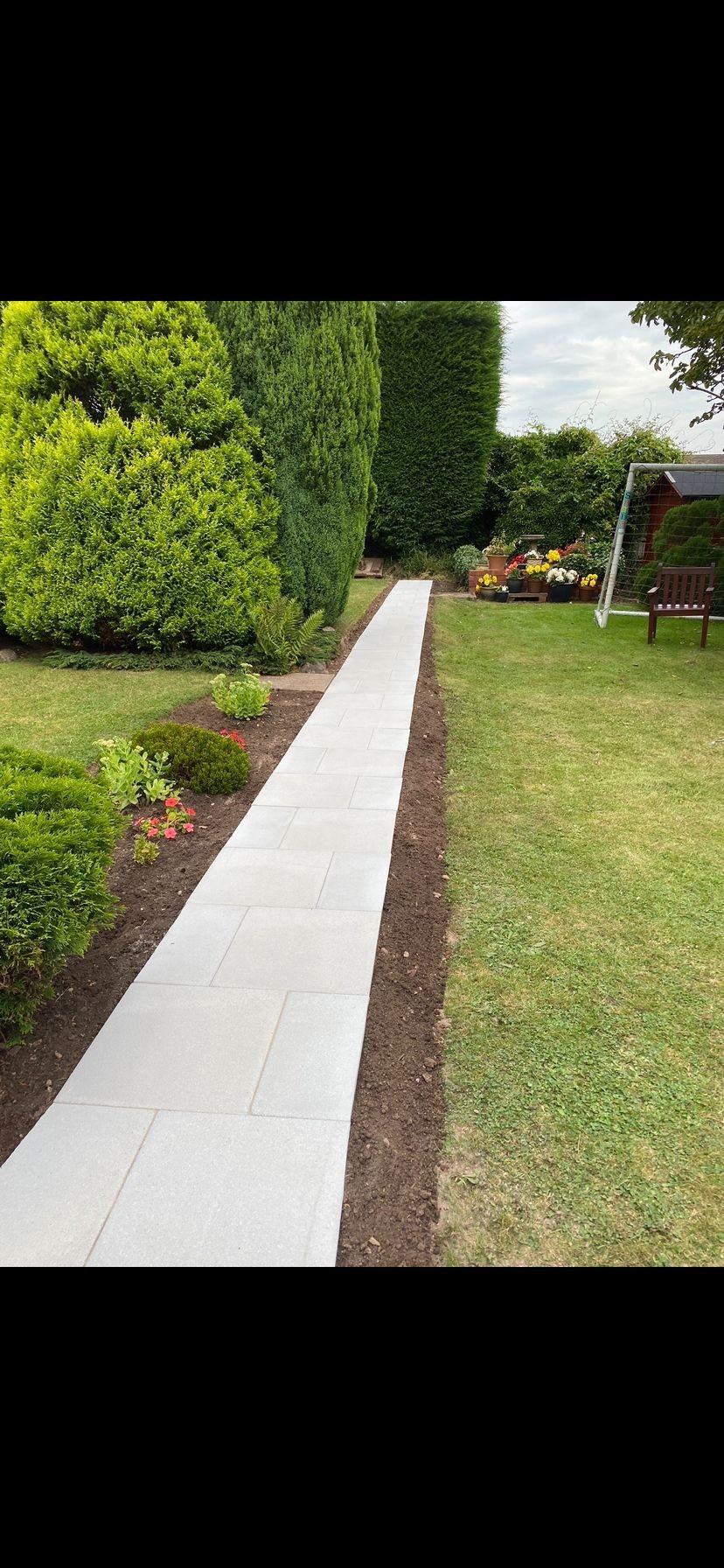 Concrete path running alongside a garden with neatly trimmed bushes, flower beds, and green grass.